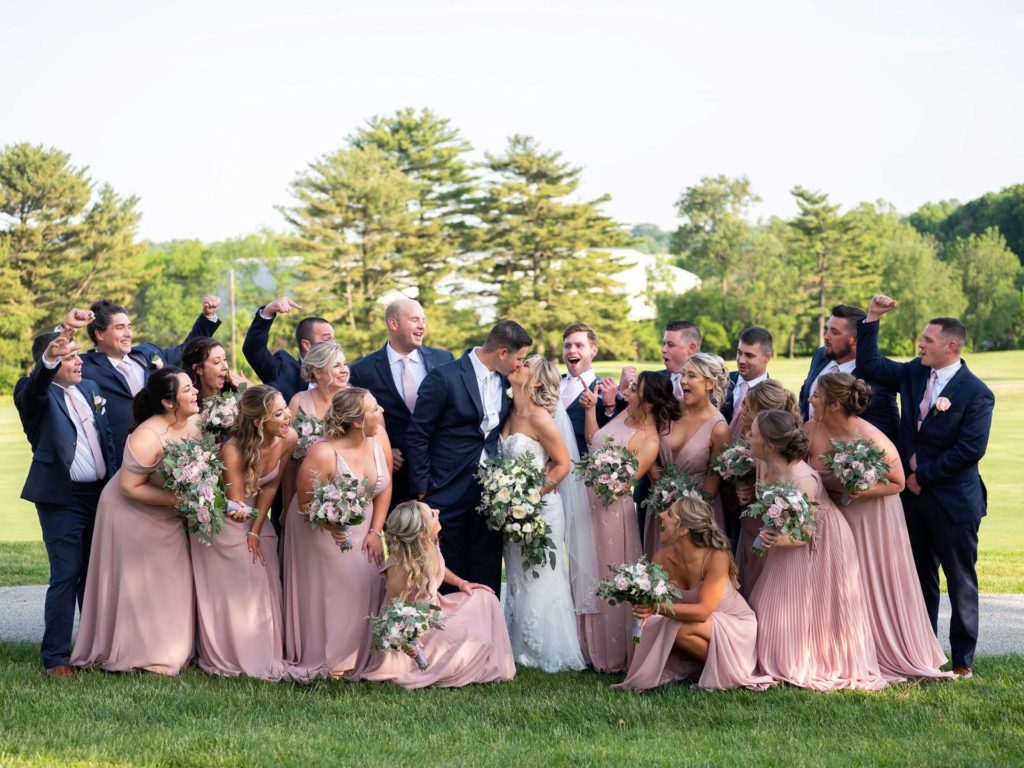 Bride and groom kissing with the bridal party surrounding them. Photographed by: Ashlee Mintz