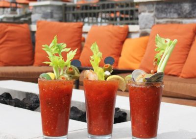 Three Bloody Marys garnished with celery, clams and shrimp with olives and lime, with firepit lounge area in background