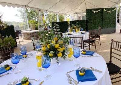 White banquet tables with lemon and greenery centerpieces under covered tent on a patio.