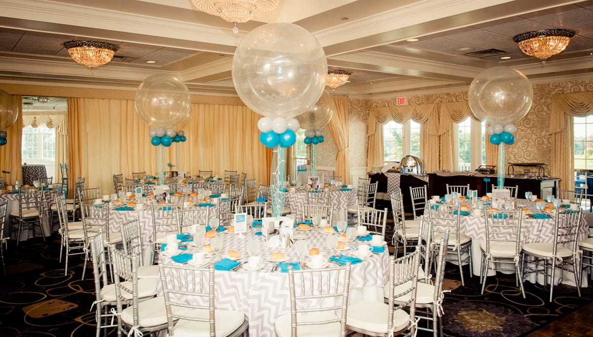 Tables set up for an event with decorative white table cloths, blue napkins, and blue, white and clear balloons as the centerpieces.  