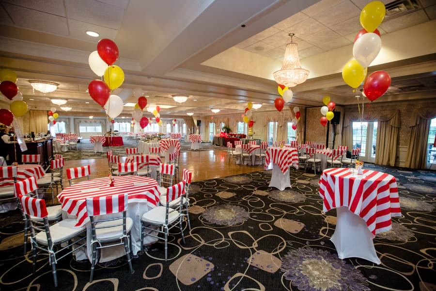 Banquet event setup with red and white striped tablecloths and chairs with red, white and yellow balloons as the centerpieces. 
