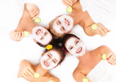 Four women laying down with the heads in a circle holding cucumbers in their hands with cold cream masks.