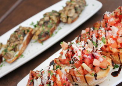 Wild mushroom bruschetta on top of crustini bread served on a white plate next to another white plate of diced tomato bruschetta served on toasted crustini bread