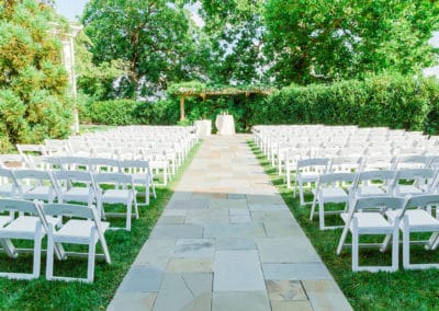 Outdoor ceremony site surrounded by greenery with white chairs on either side of the aisle and a trellis with greenery on top at the altar. Photo by Chesapeake Charm Photography