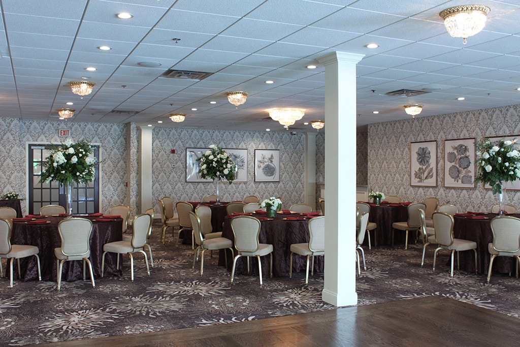 Floral patterned carpets and walls with three white columns, maroon cloth tables with gold plates and red napkins and floral centerpieces surrounded by tan and gold chairs are setup in the Fairway Ballroom.