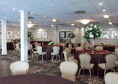 Floral patterned carpets and walls with white pillars, maroon cloth tables with gold plates and red napkins and floral centerpieces surrounded by tan and gold chairs, banquet table in left corner with chaffing dishes all in the Fairway Ballroom.