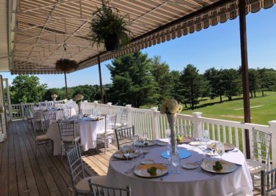 Tables set with white linen and plates on a covered veranda overlooking a golf course.