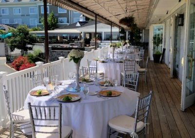 Tables set with white linen and plates on a covered veranda with restaurant in the background.