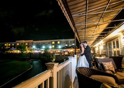 Bride and Groom standing on a covered veranda at night with brightly lit restaurant in the background.