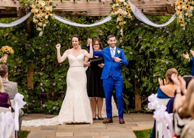 Groom holds up bride’s arm and points at her after just getting married outdoors while facing their guests at the Ceremony Garden.