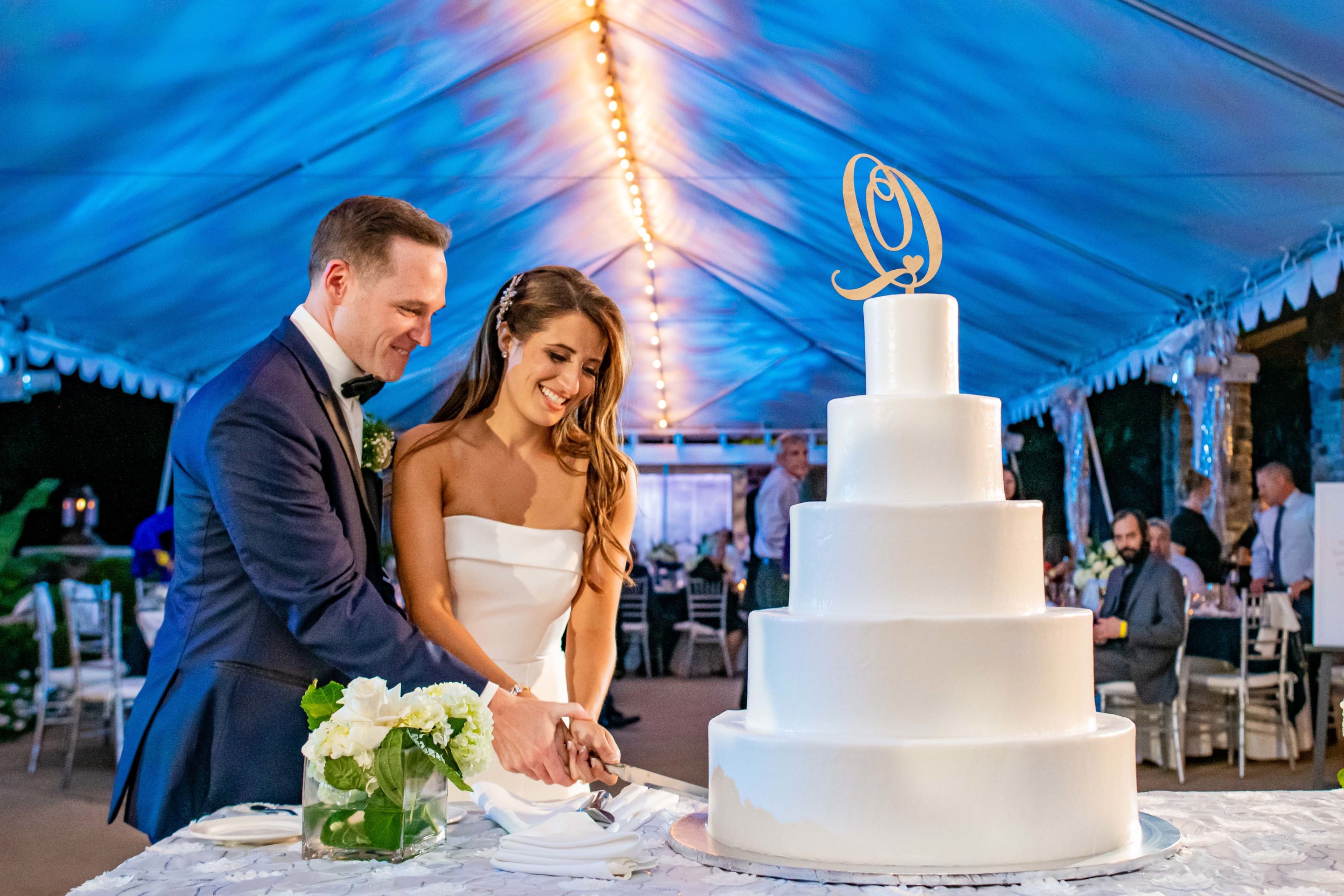 Bride and groom cutting their wedding cake under a lit canopy tent.