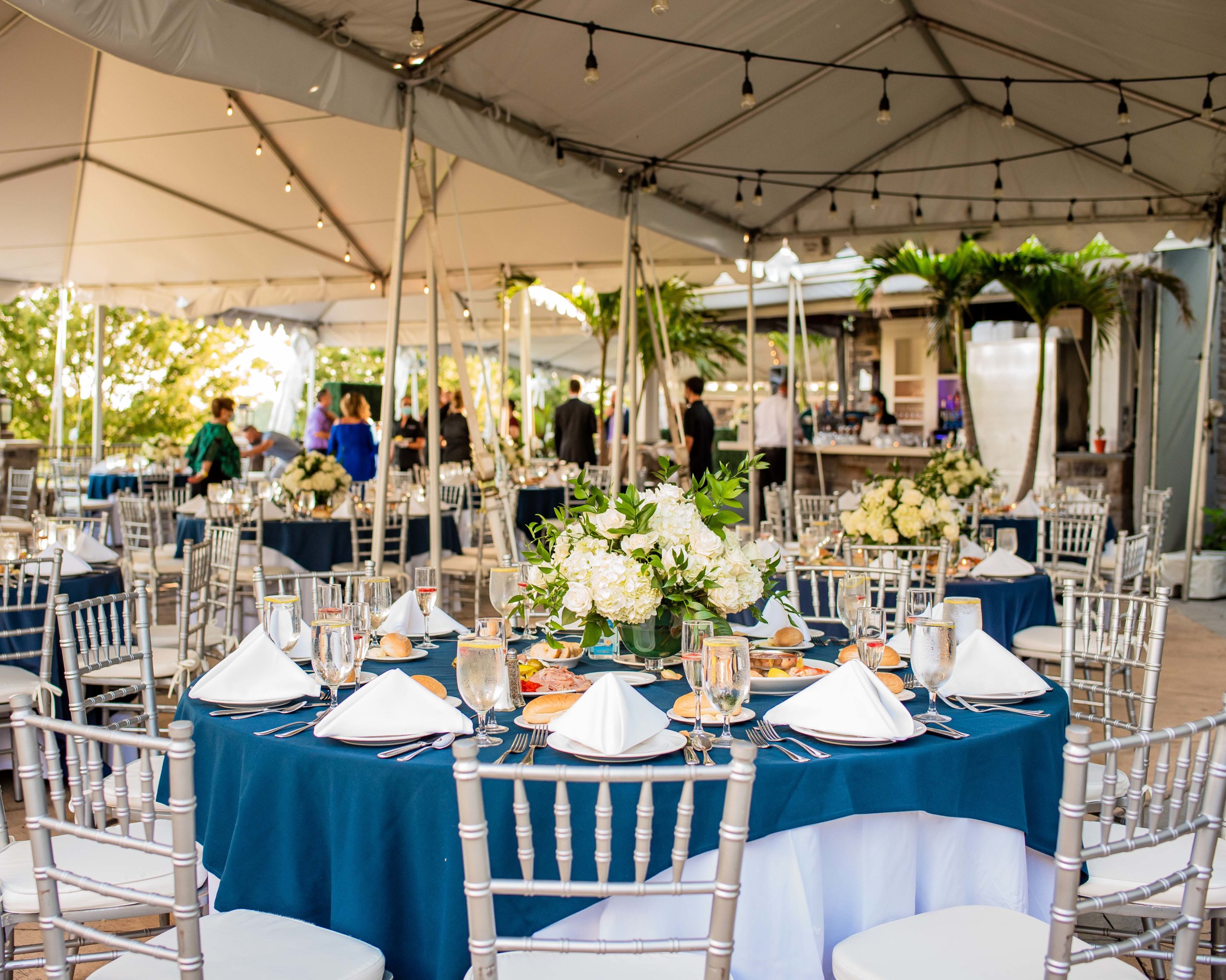 Banquet tables with blue linen and white napkins with foral centerpieces under a canopy tent with lights.