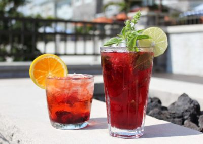 Two drinks in glasses, the left is a whiskey based drink with cherries and orange wheel in rocks glass. The drink on the right in a high ball glass is a blueberry mojito garnished with mint and a lime wheel, with a firepit in the background